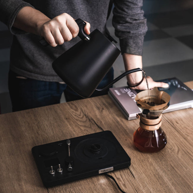 Person pouring coffee from a black kettle into a coffee dripper on a wooden table.