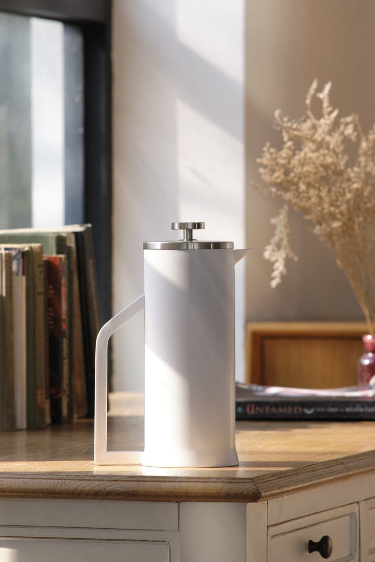 White coffee press on a wooden desk with books and decor in a bright room.