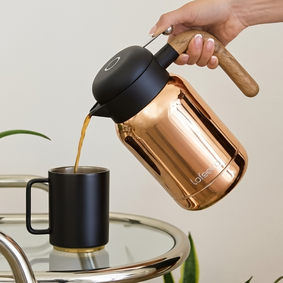 Person pouring coffee from a Lafeeca Aura copper-colored thermal carafe into a black mug on a glass table with a plant in the background.