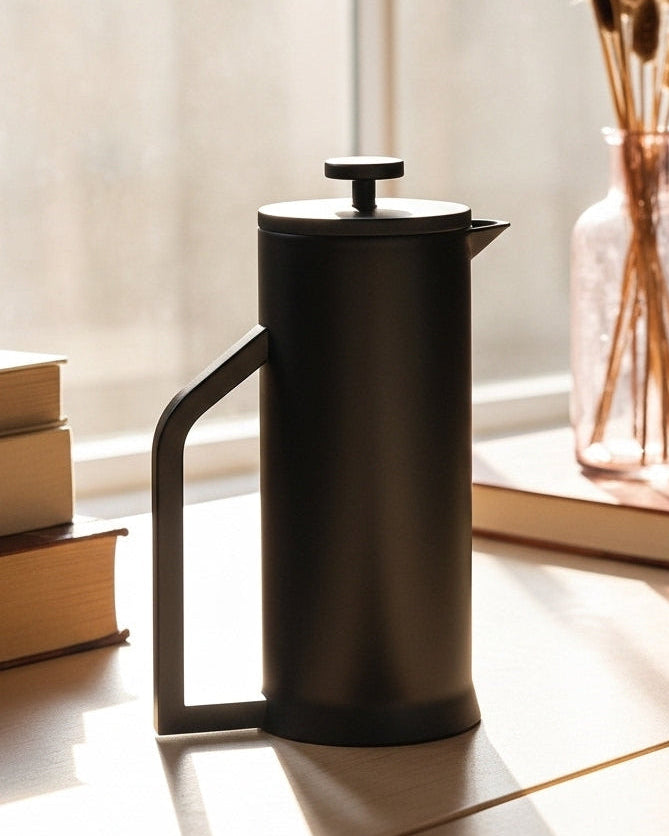 Black French press on a wooden surface with books and a vase in the background